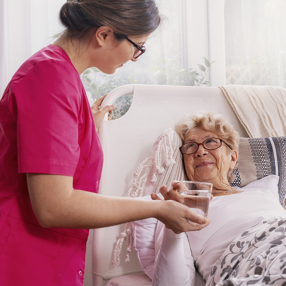Nurse in pink scrubs serves water to an elderly lady in bed.