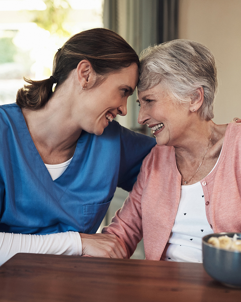 Caregiver and elderly woman smiling and leaning heads together, showing a warm and supportive bond.