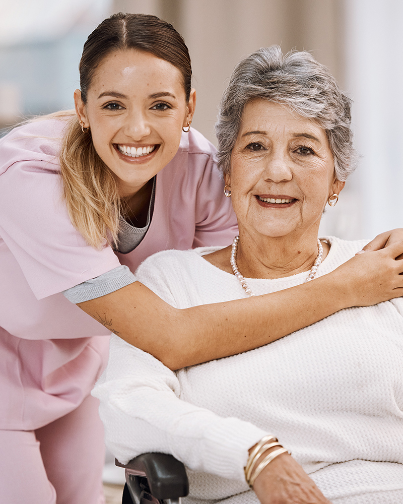 Caregiver hugging a smiling elderly woman seated in a wheelchair.